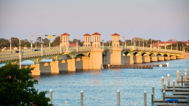 Bridge of Lions spanning blue water, with red-roofed towers, repeated arches, and marina pilings in the foreground.